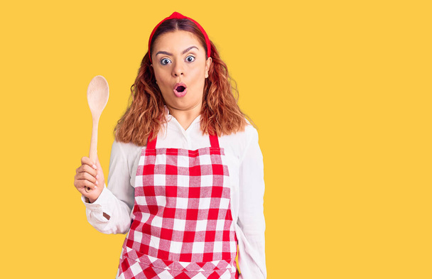 Young latin woman wearing apron holding wooden spoon scared and amazed with open mouth for surprise, disbelief face  - Zdjęcie, obraz