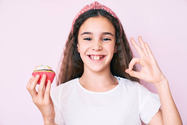 Χαριτωμένο ισπανόφωνο κορίτσι κρατώντας cupcake κάνει εντάξει υπογράψει με τα δάχτυλα, χαμογελώντας φιλικό gesturing εξαιρετικό σύμβολο  - Φωτογραφία, εικόνα