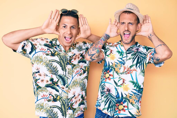 Joven pareja gay de dos hombres con sombrero de verano y camisa hawaiana sonriendo alegre jugando a echar un vistazo a un boo con las manos mostrando la cara. sorprendido y salido  - Foto, imagen