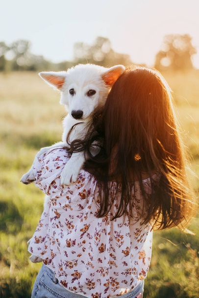 Stylová mladá žena objímající roztomilé bílé štěně v teplém západu slunce na letní louce. Šťastná dívka drží rozkošné nadýchané štěně. Krásný atmosférický okamžik. Koncepce adopce. - Fotografie, Obrázek