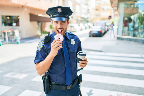 Mladý pohledný hispánský policista v policejní uniformě se šťastně usmívá. Jíst koblihy a pití šálek odnést kávu na ulici města. - Fotografie, Obrázek