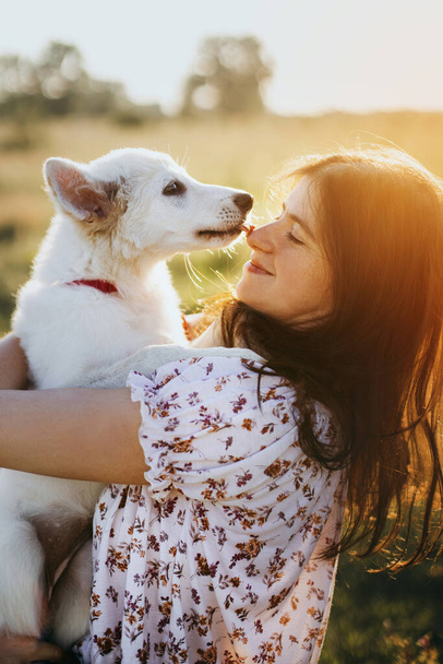 Adorable fluffy puppy kissing girl. Beautiful atmospheric moment. Stylish young woman hugging cute white puppy in warm sunset light in summer meadow. Adoption concept - Foto, immagini