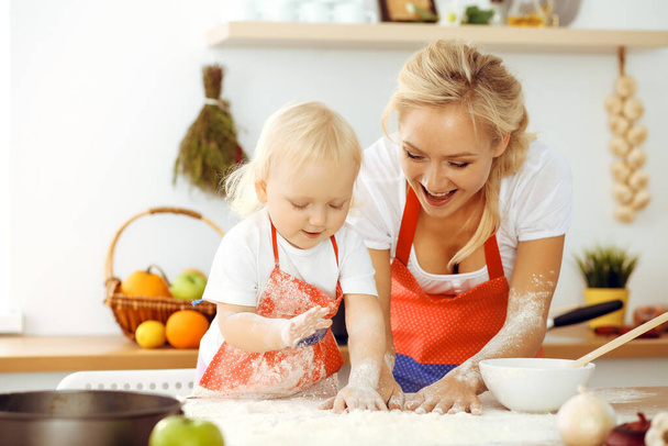 Little girl and her blonde mom in red aprons playing and laughing while kneading the dough in kitchen. Homemade pastry for bread, pizza or bake cookies. Family fun and cooking concept - Fotó, kép