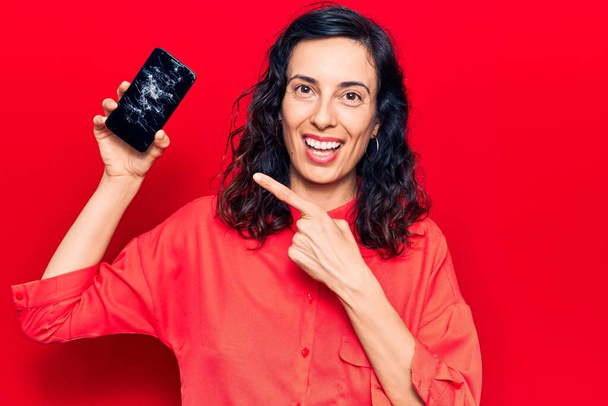 Joven mujer hispana hermosa sosteniendo teléfono inteligente roto mostrando la pantalla agrietada sonriendo feliz señalando con la mano y el dedo  - Foto, Imagen