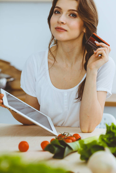 Young woman looking for a new recipe for cooking in a kitchen. Housewife is making online shopping by tablet computer and credit card - Foto, Imagen
