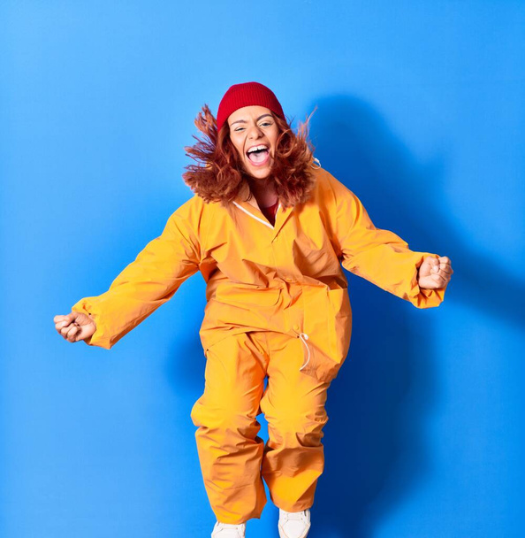 Young beautiful latin woman wearing fisher raincoat smiling happy. Jumping with smile on face celebrating with fists up over isolated blue background - Photo, Image