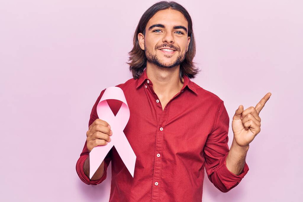 Young handsome man holding pink cancer ribbon smiling happy pointing with hand and finger to the side  - 写真・画像