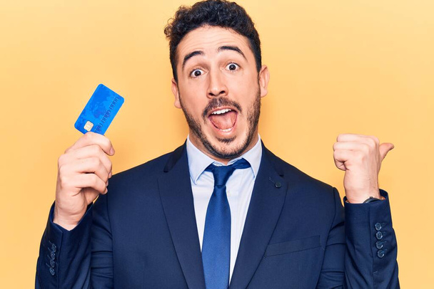 Jovem hispânico vestindo terno segurando cartão de crédito apontando polegar até o lado sorrindo feliz com a boca aberta  - Foto, Imagem