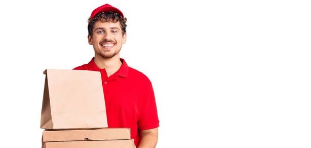 Joven hombre guapo con pelo rizado sosteniendo la caja de pizza de entrega que se ve positivo y feliz de pie y sonriendo con una sonrisa confiada mostrando los dientes  - Foto, imagen