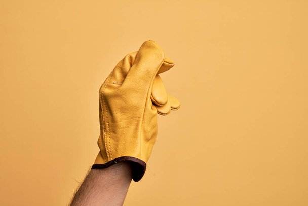 Hand eines kaukasischen jungen Mannes mit Gärtnerhandschuh über isoliertem gelben Hintergrund, der Leerstelle mit Daumenfinger hält, Geschäft und Werbung - Foto, Bild
