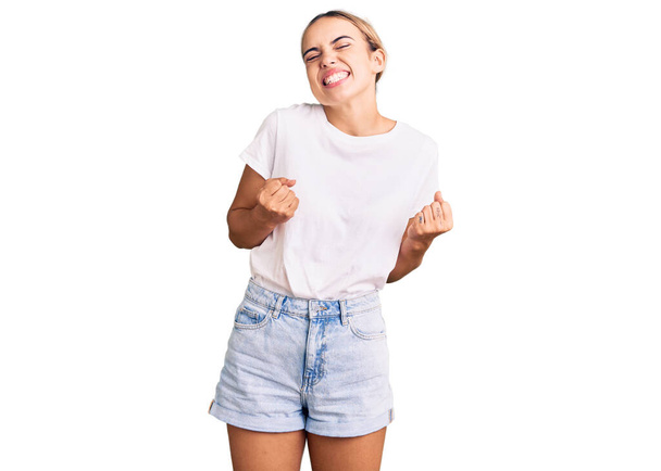 Junge schöne blonde Frau in legerem weißen T-Shirt, aufgeregt für den Erfolg mit erhobenen Armen und geschlossenen Augen, die den Sieg lächelnd feiert. Siegerkonzept.  - Foto, Bild