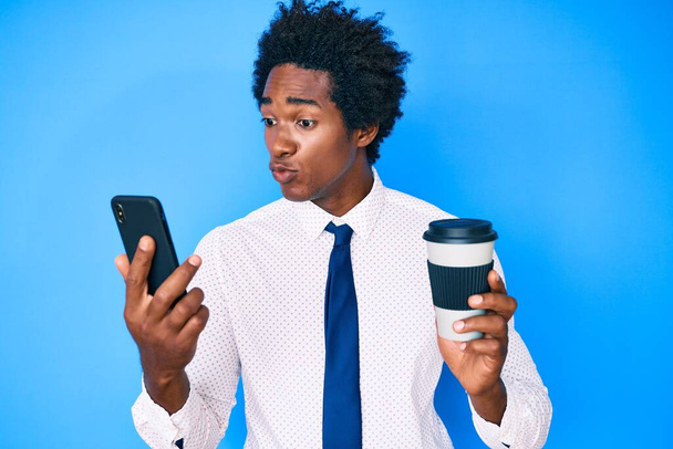 Hombre afroamericano guapo con cabello afro usando un teléfono inteligente y bebiendo una taza de café mirando a la cámara soplando un beso siendo encantador y sexy. expresión de amor.  - Foto, Imagen