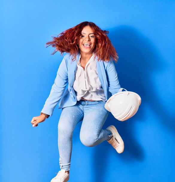 Young beautiful latin woman holding architect hardhat smiling happy. Jumping with smile on face over isolated blue background - Foto, afbeelding