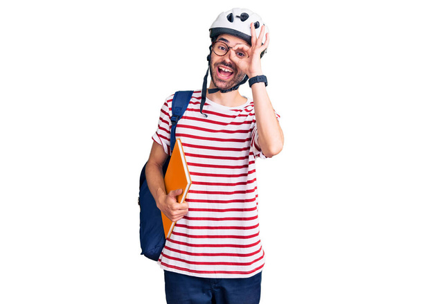 Joven hombre hispano con mochila de estudiante y casco de bicicleta sosteniendo aglutinante sonriendo feliz haciendo signo de ok con la mano en el ojo mirando a través de los dedos  - Foto, Imagen