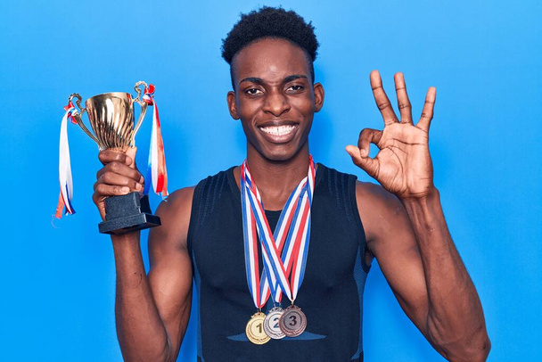 Giovane uomo afroamericano in possesso di trofeo campione indossando medaglie facendo segno ok con le dita, sorridente amichevole gesticolando eccellente simbolo  - Foto, immagini