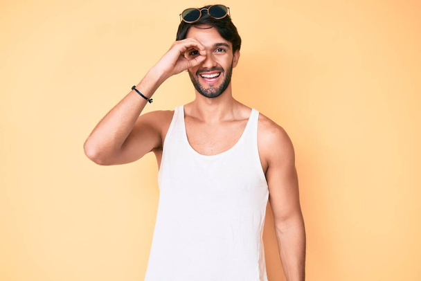 Hombre hispano guapo con mirada veraniega y gafas de sol haciendo buen gesto con la mano sonriente, ojos mirando a través de los dedos con cara feliz.  - Foto, Imagen