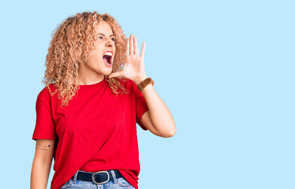 Giovane donna bionda con i capelli ricci indossa casual maglietta rossa urlando e urlando forte a lato con mano sulla bocca. concetto di comunicazione.  - Foto, immagini