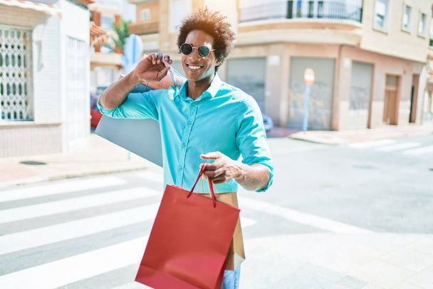 Junger gutaussehender afrikanisch-amerikanischer Mann mit Sonnenbrille, der glücklich lächelt. Mit einem Lächeln im Gesicht und Einkaufstüten in der Stadtstraße. - Foto, Bild