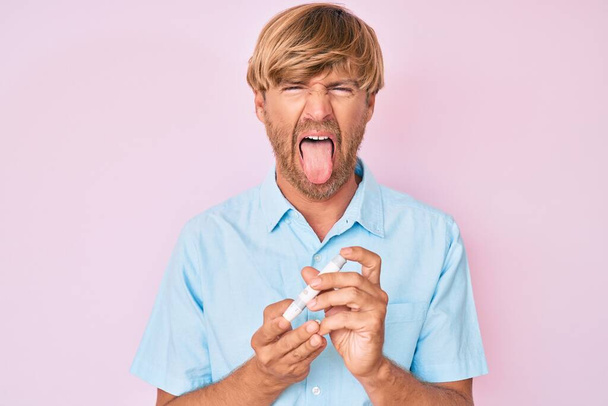 Joven hombre rubio usando glucómetro sacando la lengua feliz con expresión divertida.  - Foto, Imagen
