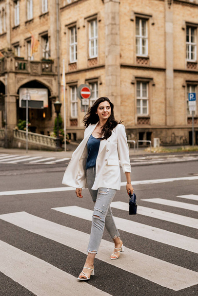 Femme attrayante souriante portant une veste de mode blanche et un jean regarde loin et traverse la rue. Une jeune femme se promène dans la ville - Photo, image