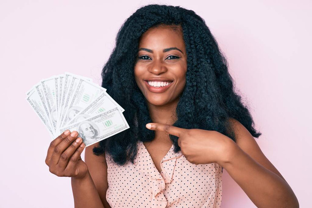 Piękna afrykańska kobieta trzyma dolary uśmiechając się szczęśliwy wskazując ręką i palcem  - Zdjęcie, obraz
