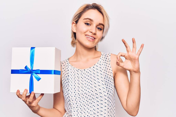Fiatal szép szőke nő gazdaság ajándék csinál rendben jel ujjakkal, mosolygós barátságos gesztus kiváló szimbólum  - Fotó, kép