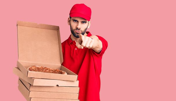 Jeune homme beau avec barbe portant des boîtes de maintien uniforme de livraison avec pizza pointant du doigt vers la caméra et à vous, geste confiant regardant sérieux  - Photo, image