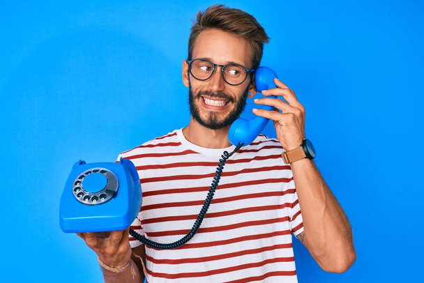 Όμορφος καυκάσιος άντρας με γενειάδα που κρατάει ένα παλιό τηλέφωνο χαμογελώντας κοιτώντας στο πλάι και κοιτάζοντας αλλού σκεπτόμενος.  - Φωτογραφία, εικόνα