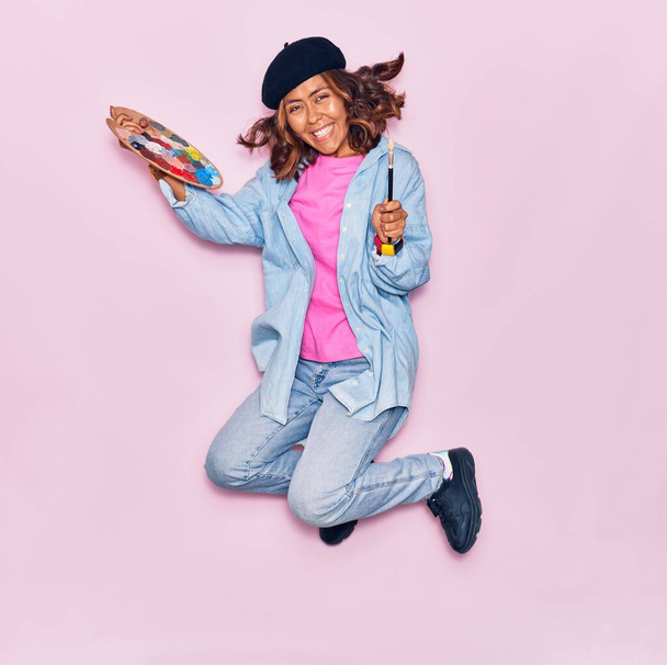 Junge schöne lateinamerikanische Künstlerin mit französischer Baskenmütze, die glücklich lächelt. Springen mit Lächeln beim Kinderschminken mit Pinsel und Palette über isoliertem rosa Hintergrund - Foto, Bild