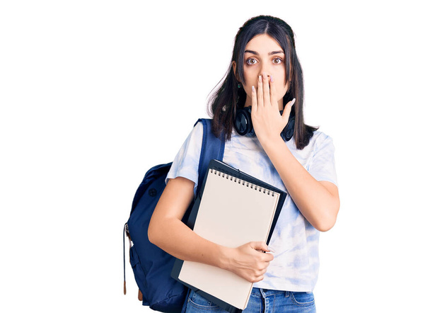 Jong mooi meisje draagt student rugzak met notitieboekje over mond met de hand, geschokt en bang voor fouten. verraste uitdrukking  - Foto, afbeelding