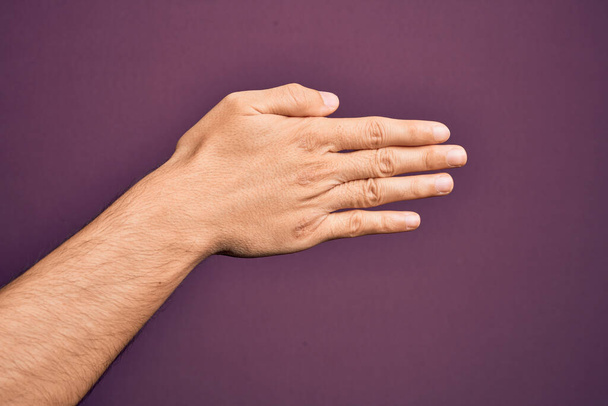 Main de jeune homme caucasien montrant les doigts sur fond violet isolé étirant et atteignant avec la main ouverte pour poignée de main, montrant le dos de la main - Photo, image