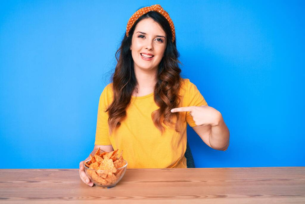 Молодая красивая брюнетка, сидящая на столе и поедающая чипсы начос, указывая пальцем на себя улыбающуюся счастливую и гордую  - Фото, изображение