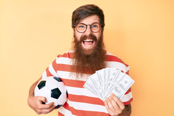 Komea nuori punainen pää mies pitkä parta tilalla jalkapallo pallo ja joukko dollareita hymyilee ja nauraa kovaa ääneen, koska hauska hullu vitsi.  - Valokuva, kuva