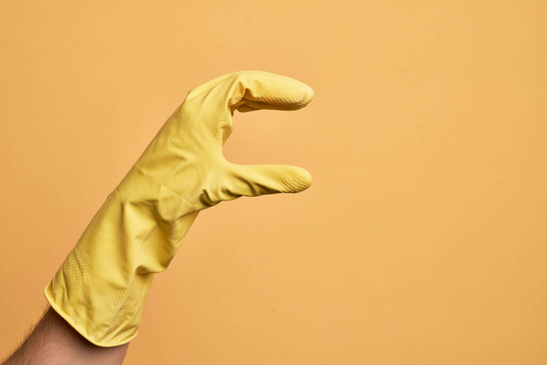 Ręka białego młodzieńca z rękawicą czyszczącą nad odizolowanym żółtym tłem zbierającym i zabierającym niewidzialną rzecz, trzymającym obiekt z palcami pokazującymi przestrzeń - Zdjęcie, obraz