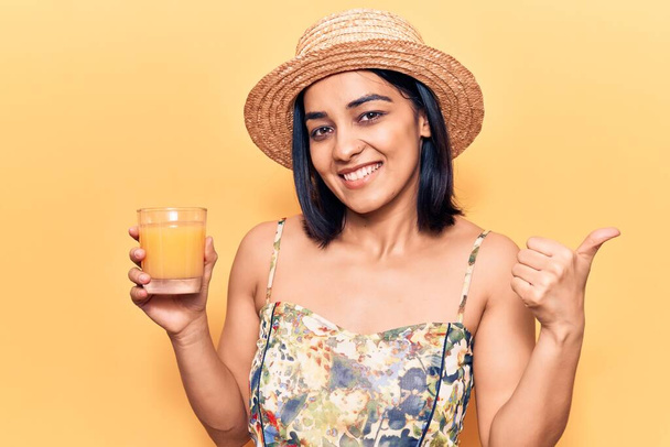 Νεαρή όμορφη Λατίνα γυναίκα φοράει καλοκαιρινό καπέλο πίνοντας ποτήρι χυμό πορτοκαλιού δείχνοντας τον αντίχειρα στο πλάι χαμογελώντας χαρούμενη με ανοιχτό στόμα  - Φωτογραφία, εικόνα