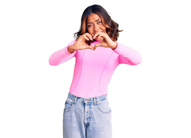 ピンクのシャツを着た若い美しい混合人種の女性は、ハートのシンボルと手で形を示す愛に微笑んでいます。ロマンチックなコンセプト.  - 写真・画像