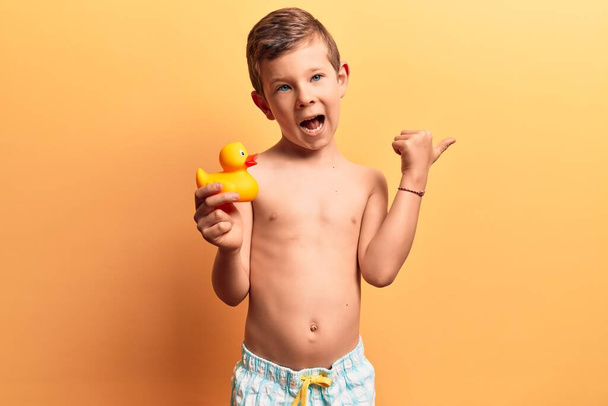 Lindo niño rubio con traje de baño sosteniendo el juguete del pato señalando el pulgar hacia el lado sonriendo feliz con la boca abierta  - Foto, imagen