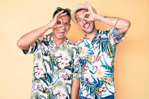 Jeune couple gay de deux hommes portant un chapeau d'été et une chemise hawaïenne faisant un geste correct comme des jumelles sortant la langue, les yeux regardant à travers les doigts. expression folle.  - Photo, image