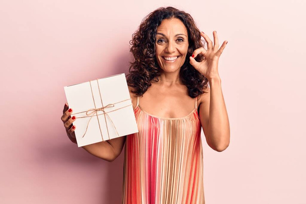 Middle age beautiful woman holding gift doing ok sign with fingers, smiling friendly gesturing excellent symbol  - Foto, Imagen