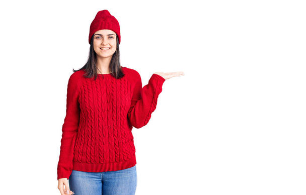 Młoda piękna dziewczyna ubrana w sweter i wełnianą czapkę uśmiechnięta radosna prezentacja i wskazująca dłonią na aparat fotograficzny.  - Zdjęcie, obraz