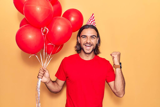 Young handsome man holding balloons screaming proud, celebrating victory and success very excited with raised arms  - Photo, Image