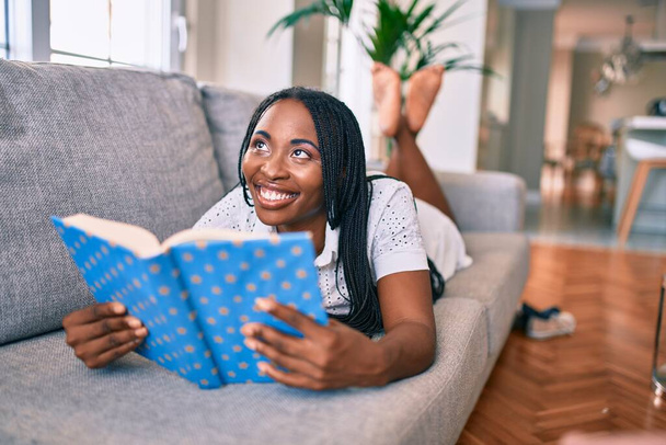 Joven mujer afroamericana sonriendo feliz tendida en el libro de lectura del sofá en casa - Foto, Imagen