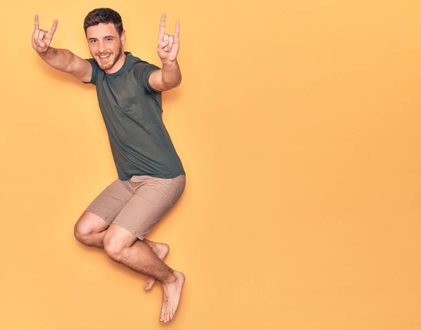 若いハンサムなヒスパニック系の男性が幸せな笑顔カジュアルな服を着ている。独立した黄色の背景の上に指で角記号を行う顔に笑顔でジャンプ - 写真・画像
