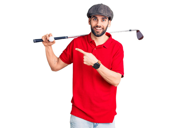 Joven hombre guapo con barba jugando al golf sosteniendo el palo y la pelota sonriendo feliz señalando con la mano y el dedo  - Foto, imagen