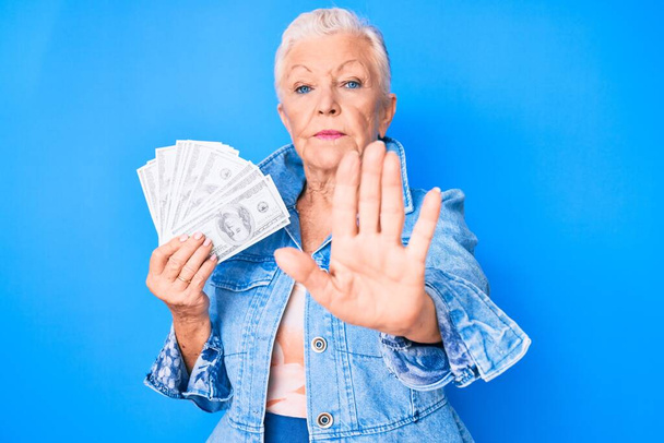Старшая красивая женщина с голубыми глазами и седыми волосами держа доллары с открытой рукой делает знак стоп с серьезным и уверенным выражением лица, защитный жест  - Фото, изображение