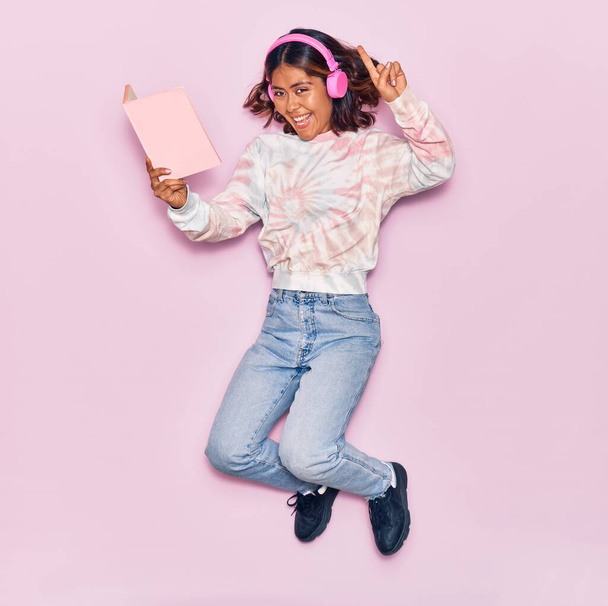 Young beautiful latin woman listening to music using headphones smiling happy. Jumping with smile on face reading book over isolated pink background - Photo, image