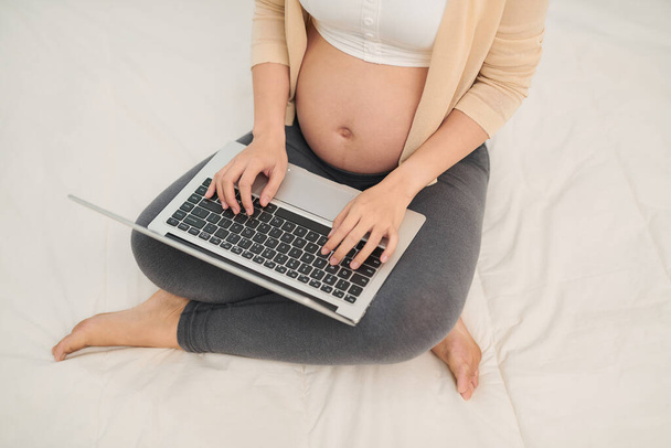 Femme enceinte en vêtements blancs assis et utilisant un ordinateur portable au lit. - Photo, image