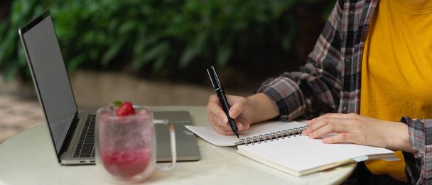 Вид сбоку женщины-фрилансера, делающей запись в ноутбуке во время работы с макетом ноутбука на столе в саду  - Фото, изображение