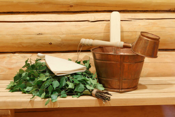 Μια φρέσκια σκούπα σημύδας και χάλκινη δεξαμενή και κουτάλα είναι σε ένα ξύλινο πάγκο στη σάουνα. - Φωτογραφία, εικόνα