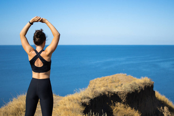 Visszapillantás a sportos fiatal barna nő nyújtózkodó karok edzés előtt futás, álló sziklafal mellett kék tenger a napsütéses napon. Reggeli testmozgás a szabadban természet táj. - Fotó, kép